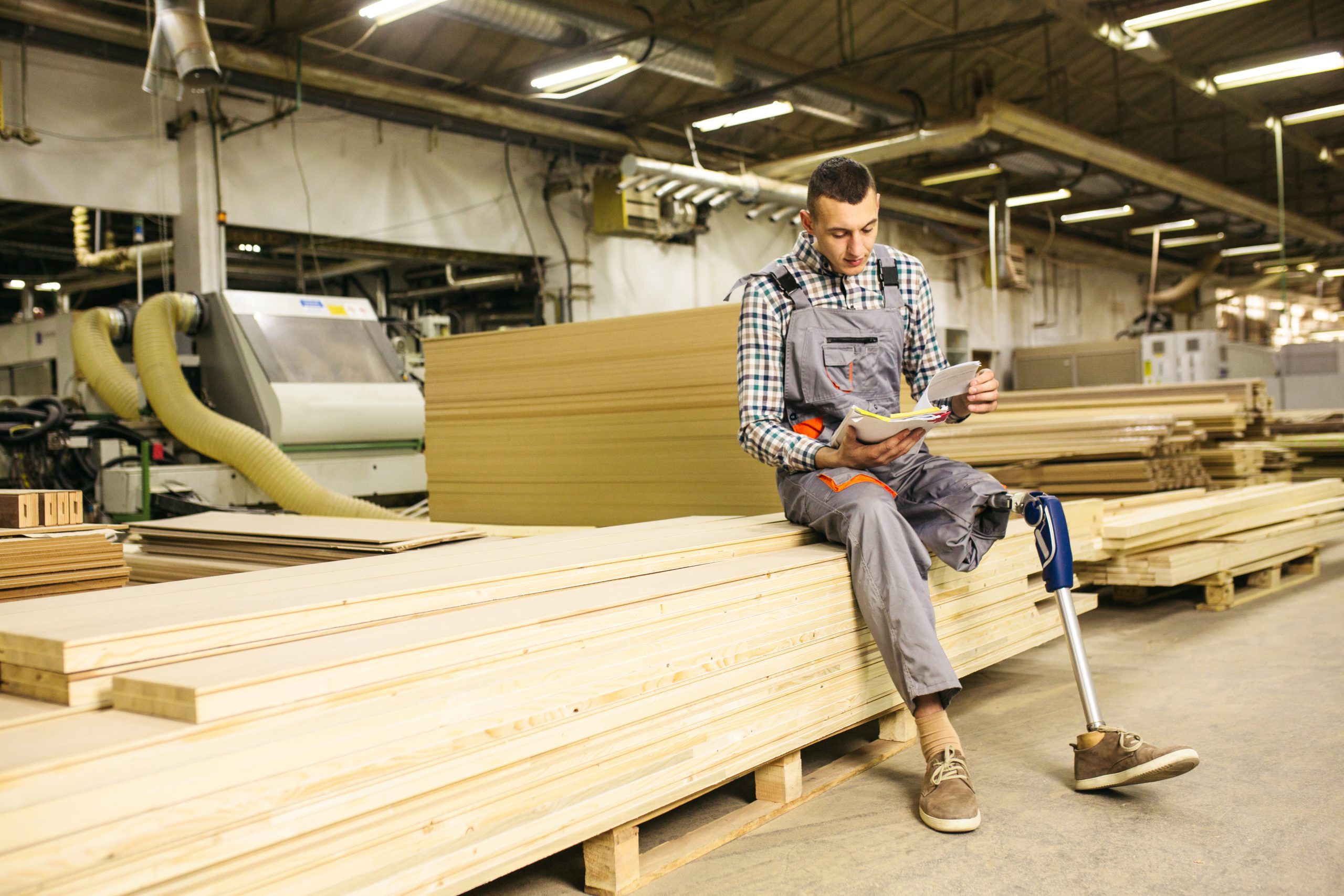 Young carpenter with prosthetic leg at the factory lifting wood plank